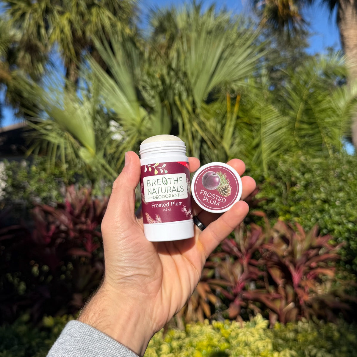 Hand holding a bottle labeled 'Breathe Naturals Frosted Plum' against a tropical background person using all natural non-toxic deodorant