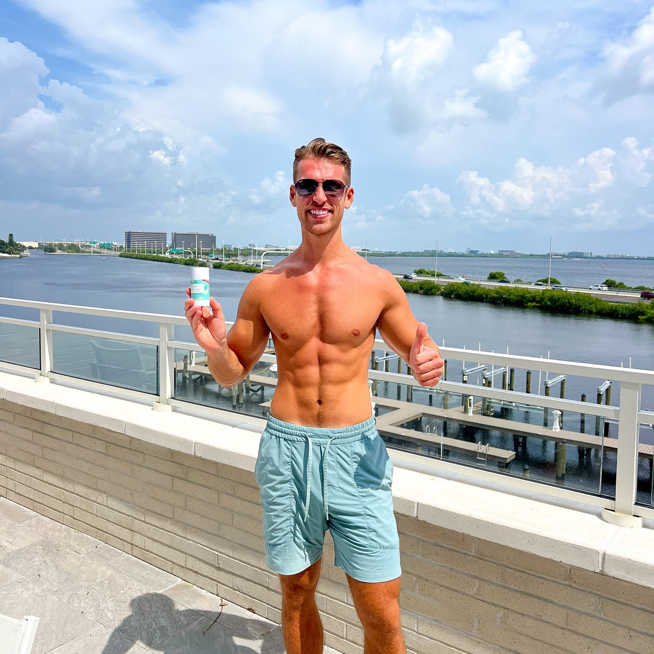 Man in light blue shorts holding a water bottle on a rooftop with a waterfront view.