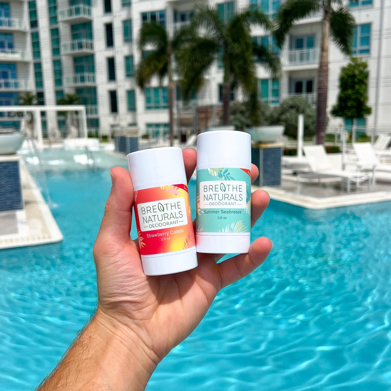 Two Breathe Naturals products held in front of a pool with buildings and palm trees in the background